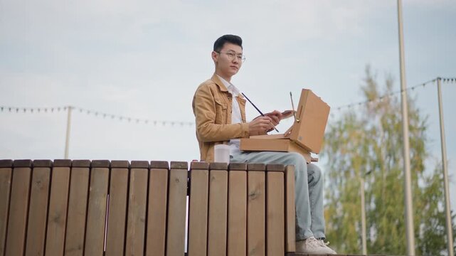 asian man painting on wooden deck, portable box easel and sketch supplies, overcast sky with string lights, autumn trees in background, casual jacket, contemplative atmosphere, urban street art vibe