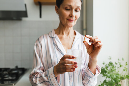 Daily routine of middle-aged healthy and good-looking female for collagen and antioxidant support based on taking dietary supplements, woman holding gel capsule of fish oil and glass of water