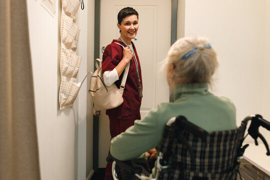 Indoor picture of kinesiotherapist leaving place of her patient in wheelchair with her back turned to camera, saying bye after finishing her therapy for today, standing at front door with backpack