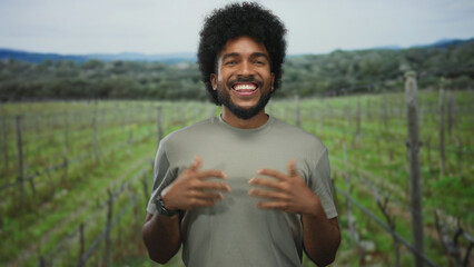 Fototapeta premium Man with afro smiling in a lush green vineyard giving thumbs up in a park setting.