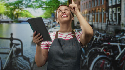 Woman holding tablet points finger up on street in amsterdam wearing apron and striped shirt  optimism learning. © Krakenimages.com