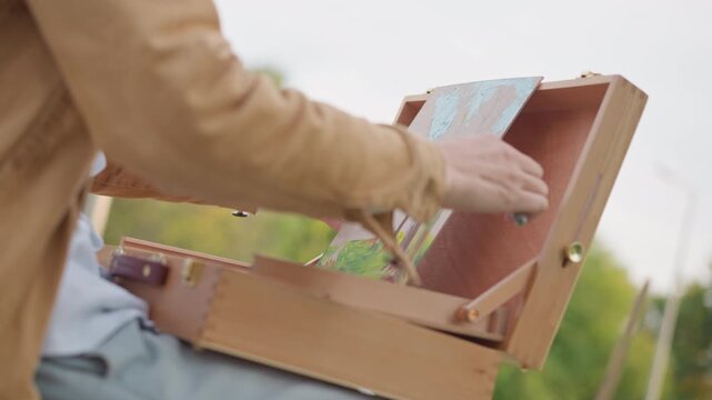 artist setting up plein air easel, jacketed painter leans into wooden box, transferring sketch into color study, park backdrop with green trees and soft sunlight, relaxed slow living session