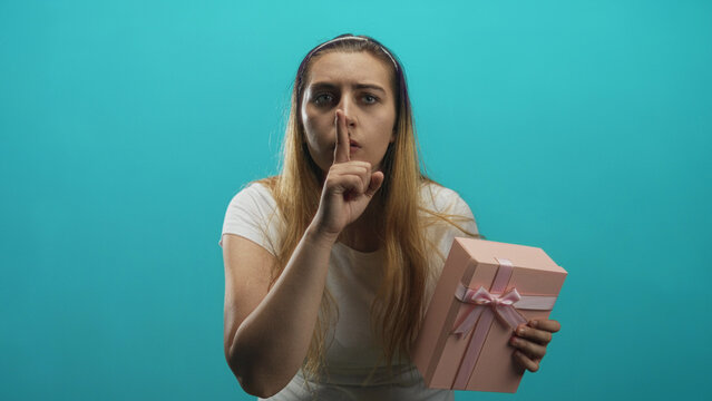 Woman holding a pink present and pressing finger to lips in studio, leaning forward with worried glance and tense expression; secret silence.