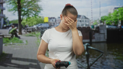 Woman in white t shirt holding a black gamepad and covering face with right hand on a street by a canal in daylight  gaming frustration. © Krakenimages.com