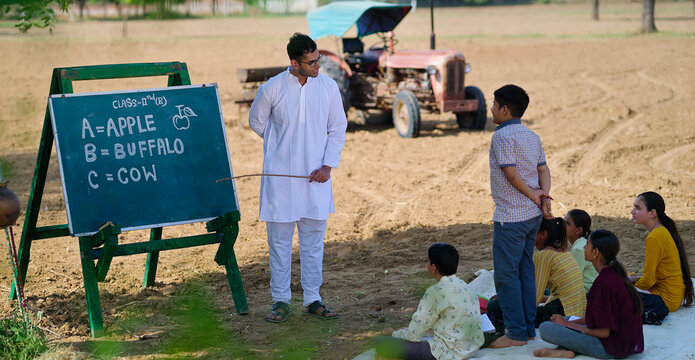 Indian young adult gen z male man guy talk speak teach to villager child day under tree rural area. Happy girl boy group sitting on mat do study look board learn read English at open class field place