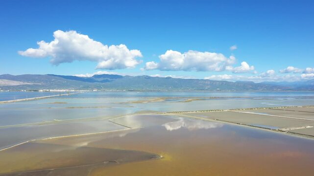 Aerial view of salt pans and shallow water reflecting clouds at Marais salant de Missolonghi, Greece, with distant mountains under a clear blue sky.