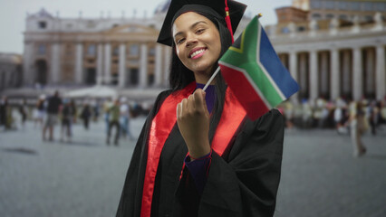 Fototapeta premium Young woman waves south african flag while wearing black graduation gown and cap in front of vatican building; pride accomplishment celebration.