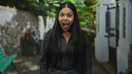 Obraz premium Young latin woman holds face with hands on cheeks in street beside stone wall with blurred bench; surprise.