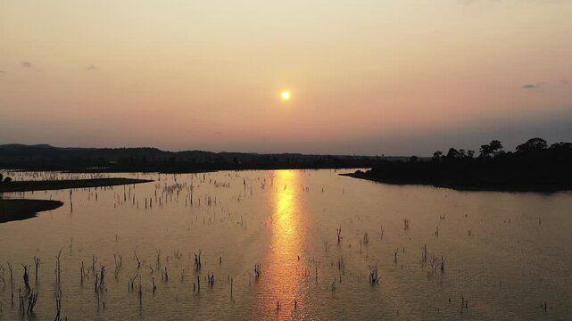 Tranquil sunset view over Lac de Nakai, with the sun reflecting on calm water and silhouettes of tree stumps emerging from the lake. Serene natural landscape with distant hills and a peaceful