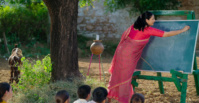 Indian mature adult teacher do study work teach to villager young girl boy rural area child group sitting on mat look board learn English desi life open class day time field ground place