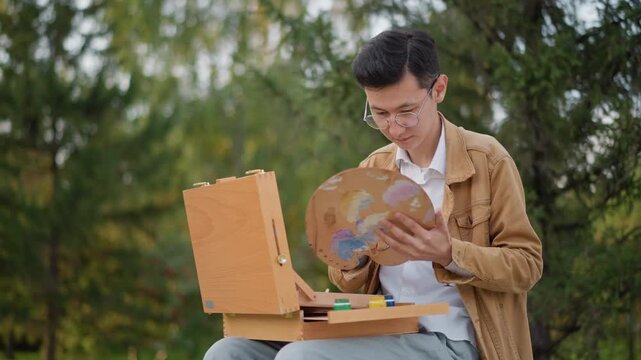 focused asian male mixing colors outdoors at easel and paint box, freelance illustrator studying hues and brushwork, closeup of hands and wooden palette, calm park backdrop with trees and natural