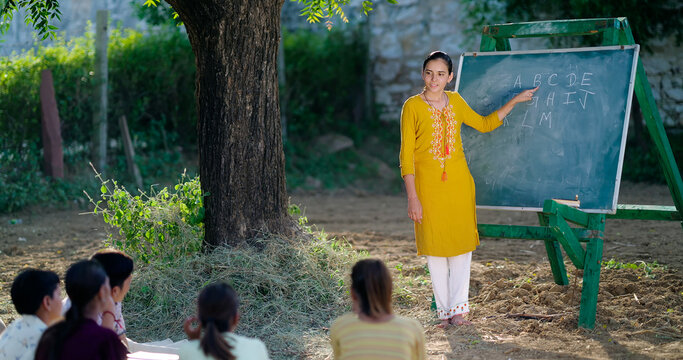 Indian adult gen z female tutor standing teach English to villager girl boy outdoor rural area. Happy child group sitting on mat look board learn do study new day time outside ground place