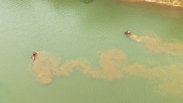 Aerial view of two elephants with riders bathing and stirring up sediment in a calm river, likely at an elephant village. Natural, tranquil scene with green water and visible water movement.