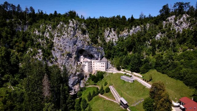 Aerial view of Predjama Castle nestled within a gaping cave mouth in a steep, lush, green cliffside contrasted by the castle's pale stone, Postojna, Slovenia.