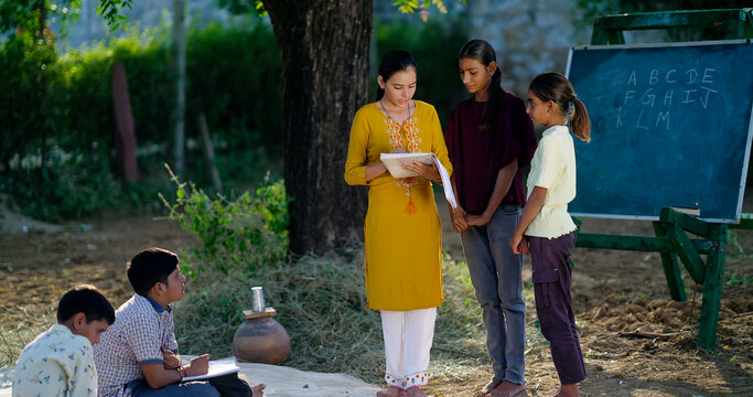 Indian young adult female hold book do study helping to read teen age girl class rural area place. Boy student group talk speak sitting on mat enjoy spend break time have fun joy open air ground