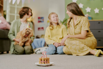 Obraz premium Caucasian child sitting between two Caucasian women on floor, smiling and holding hands, small dog with party hat sitting nearby, birthday cake with number three candle in foreground