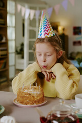 Fototapeta premium Vertical of Caucasian girl child wearing party hat blowing out candles on birthday cake while sitting at table indoors, hands resting under chin, celebrating birthday in home setting