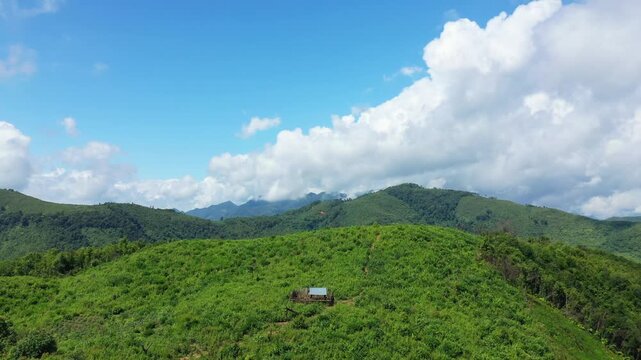 Lush green hills and distant mountains under a bright blue sky in the countryside between Phou Khoun and Luang Prabang, Laos. A small isolated hut sits atop the hill, surrounded by natural vegetation.