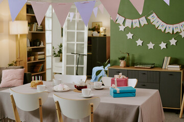 Fototapeta premium Birthday party table setting featuring cake with candles, wrapped gifts, teacups, and pastries arranged on table, pastel bunting and happy birthday banner decorating background wall
