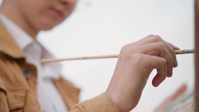 closeup hands holding paintbrush at easel, street sketcher wearing brown jacket focuses on delicate strokes on canvas and palette, soft bokeh city background, calm pleinair atmosphere, intermittent