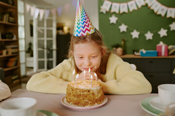 Fototapeta premium Portrait of Caucasian girl child wearing party hat leaning forward blowing out candles on birthday cake during celebration at home, smiling and appearing joyful with cake in foreground