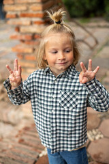 A close-up portrait of a child in a plaid shirt showing a peace sign with a playful smile, with brick wall background