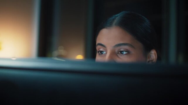Close face view of Asian businesswoman looking at computer screen. Adult female focusing on monitor. Sitting indoors at night office. Concentrating on digital work tasks. Professional mood.