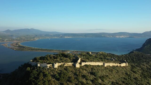 Aerial view of the historic Navarino Castle surrounded by lush greenery with a scenic bay and distant mountains under a clear blue sky. The ancient stone fortress stands prominently on a hilltop