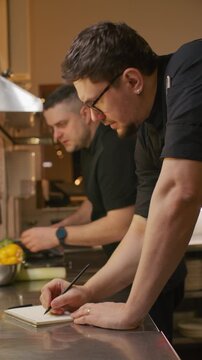 Male chef writing notes in notebook and discussing menu with colleague while standing at kitchen counter in restaurant kitchen. Vertical format footage