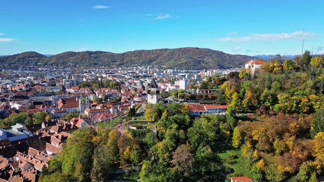 Aerial view of the vibrant city of Graz with its terracotta rooftops, contrasted by the lush green Schlossberg hill under a clear blue sky, Graz, Styria, Austria.