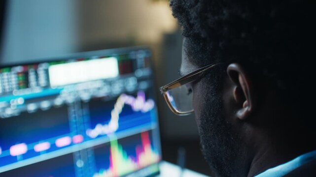 Over shoulder view of African American man watching stock charts on monitor. Adult male wearing glasses. Analyzing market data on screen. Sitting indoors at night office. Focused trading session.