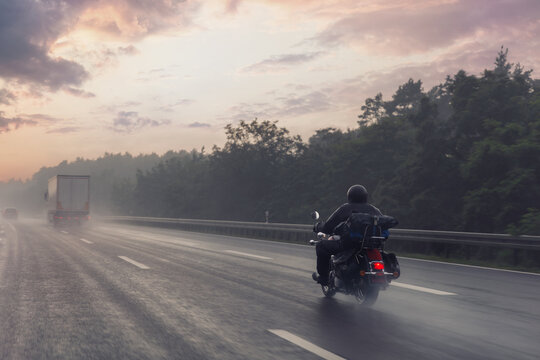 Motorcycle rider speeds along misty highway sunrise red tail light glow rain soaked asphalt drifting spray. Forest silhouette frames horizon truck fades fog evoking freedom adventure, risk solitude