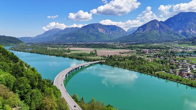 Aerial view of the striking turquoise River Drau, crossed by a modern bridge amidst verdant hills and distant mountains, Ferlach, Carinthia, Austria.