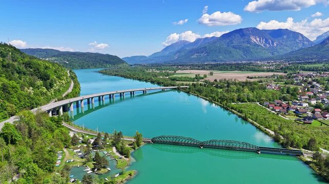 Aerial view of bridges spanning across the turquoise River Drau, framed by verdant hills and distant mountains, Ferlach, Carinthia, Austria.