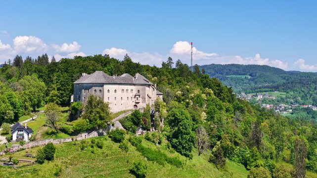 Aerial view of a historic castle on a lush green hill surrounded by a dense forest with a radio tower in the background, Ferlach, Carinthia, Austria.