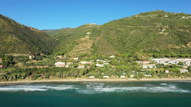 Aerial view of the coastline and hillside village of Acciaroli, Italy, with lush greenery and gentle waves along the shore under a clear blue sky.