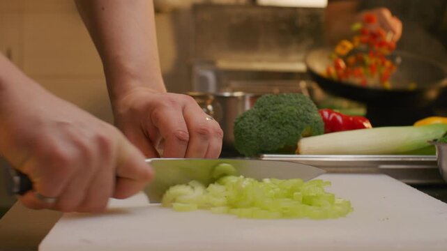 Close up shot of chef slicing fresh celery with knife on cutting board in professional restaurant kitchen with bright flambe flame burning in background during cooking process