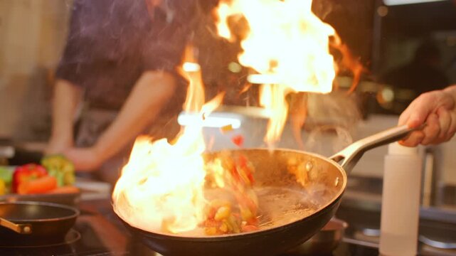 Chef igniting fire with culinary torch and flambe cooking vegetables in frying pan on hot stove as large flame rising during meal preparation in professional restaurant kitchen. Close up view