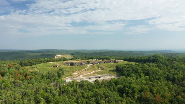 Drone shot of Fort de Douaumont surrounded by lush green forest and rolling hills under a partly cloudy sky in France.