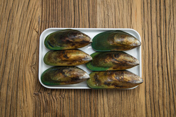 Six glossy green mussels are lined up on a white tray placed over a textured wooden board. The shells show natural color variation, presenting a clean seafood ingredient ready for cooking.