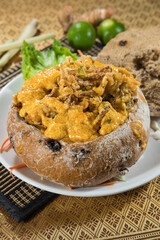 A hearty curry fills a rustic round bread bowl, topped with crispy shallot. Lemongrass, lime and leafy garnish on the table hint at fragrant spices and a comforting, shareable dish.