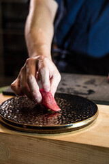 At a sushi counter, a chef places a bright tuna nigiri onto a textured black plate. The tight framing and soft light emphasize the color of the fish and the refined service moment.