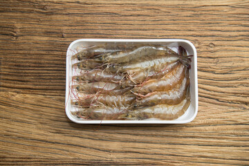 A white plastic tray holds a tidy row of raw shrimp with shells and antennae. Moist translucent bodies suggest freshness, photographed overhead on textured wood.