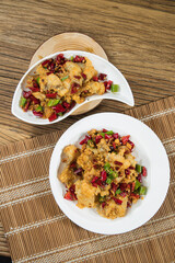 Two plates present crispy chili chicken tossed with dried red pepper, green pepper and aromatics. The overhead view on bamboo mats shows bold color, heat and a lively sharing style dish.