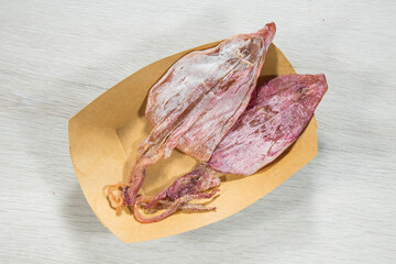 Two pieces of dried squid rest in a brown paper tray on a light table. The overhead angle shows the natural texture and powdery surface, suggesting a classic market style snack ingredient.