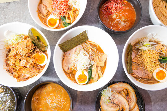 Several ramen bowls fill the frame, each with noodles, chashu, soft eggs, seaweed and varied broths. The bright overhead view showcases variety, warmth and the comfort of a noodle shop experience.