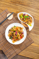 Two plates of stir fried meat and vegetables sit on bamboo mats. Slices of pepper, onion and carrot add color and gloss, suggesting a lively family style dish made for sharing.