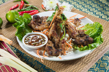 A plate of grilled lamb chops stands over crisp shredded vegetables and lettuce with a small dipping sauce on the side. Red chili and limes decorate the patterned table for a lively look.