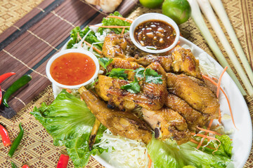 A generous platter of grilled chicken pieces sits over shredded salad greens with two dipping sauces. Cilantro, chili and lime add freshness, styled on patterned cloth with bamboo mat accents.