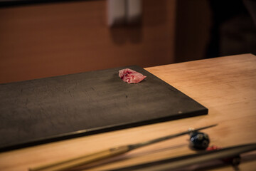 A single slice of fresh sashimi rests on a dark board at a wooden counter. Soft focus tools lie nearby, framing a quiet preparation moment at a sushi bar.